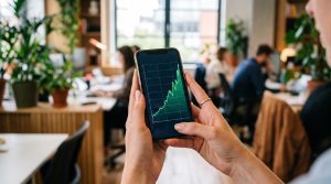 Como montar um plano de aposentadoria para autônomos em etapas práticas 3 Mãos segurando smartphone com aplicativo de investimentos aberto, mostrando gráfico de crescimento patrimonial no longo prazo, fundo desfocado de ambi